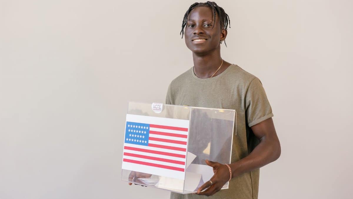 Smiling young adult holding a ballot box with an American flag, emphasizing the importance of voting.