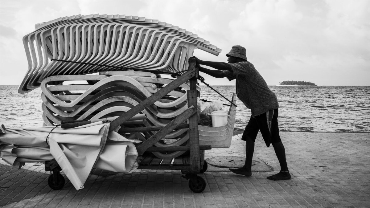 A man pushes stacked beach chairs by the seaside in a black and white photograph.