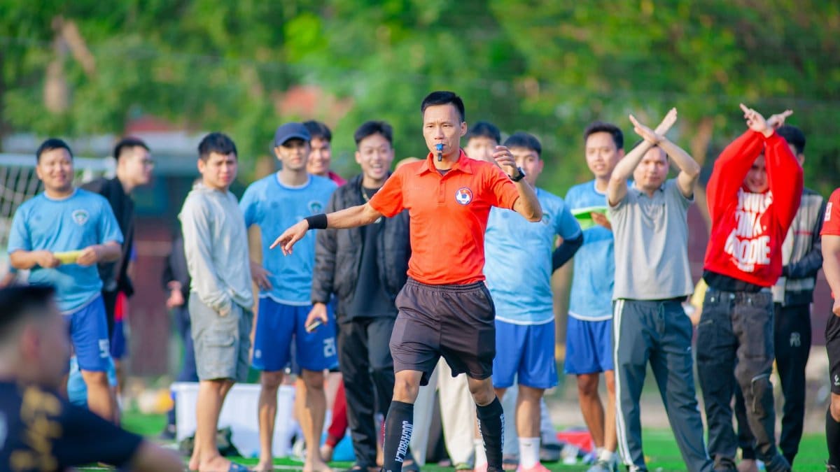 A referee in orange signals during a soccer game with players watching in the background.
