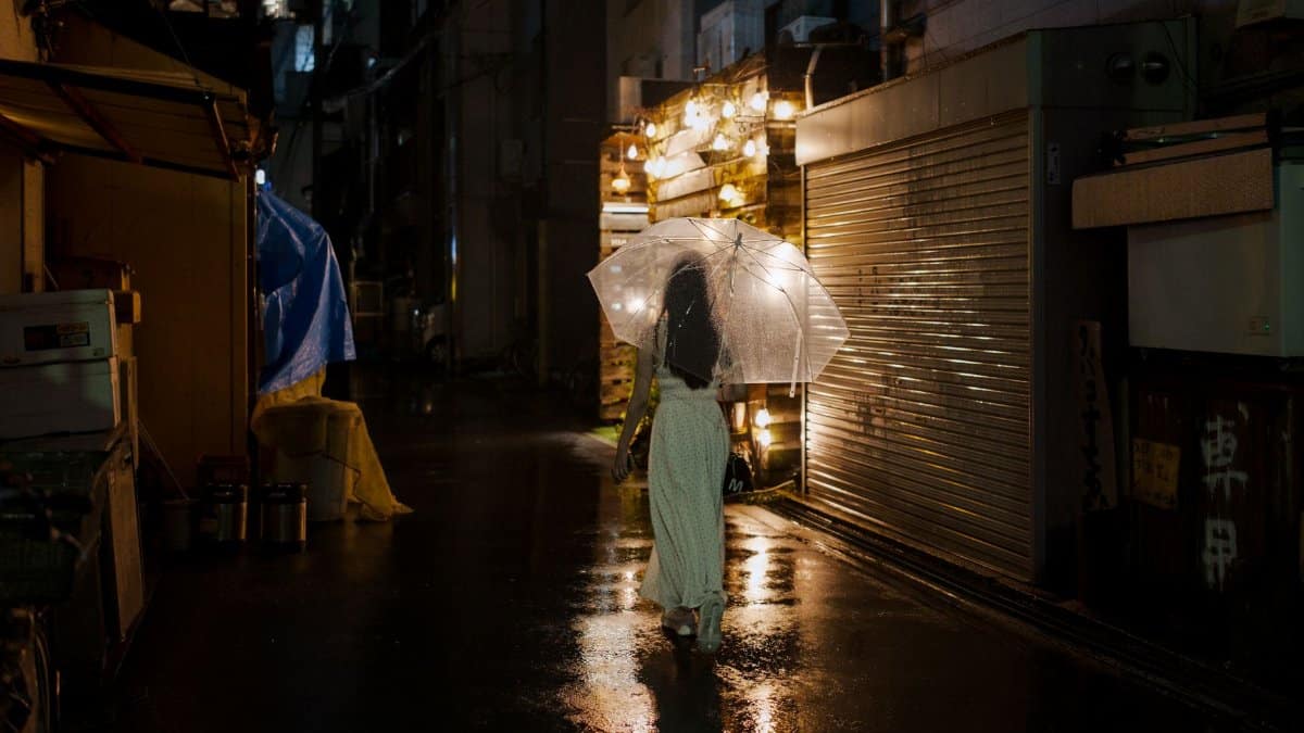 A woman walks under an umbrella in a rainy alley lit by street lights in Osaka, Japan.