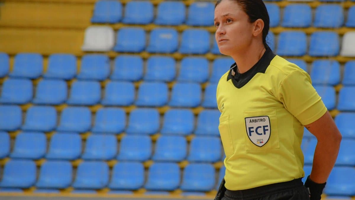 Woman futsal referee in yellow uniform stands focused in empty indoor stadium with blue seats.