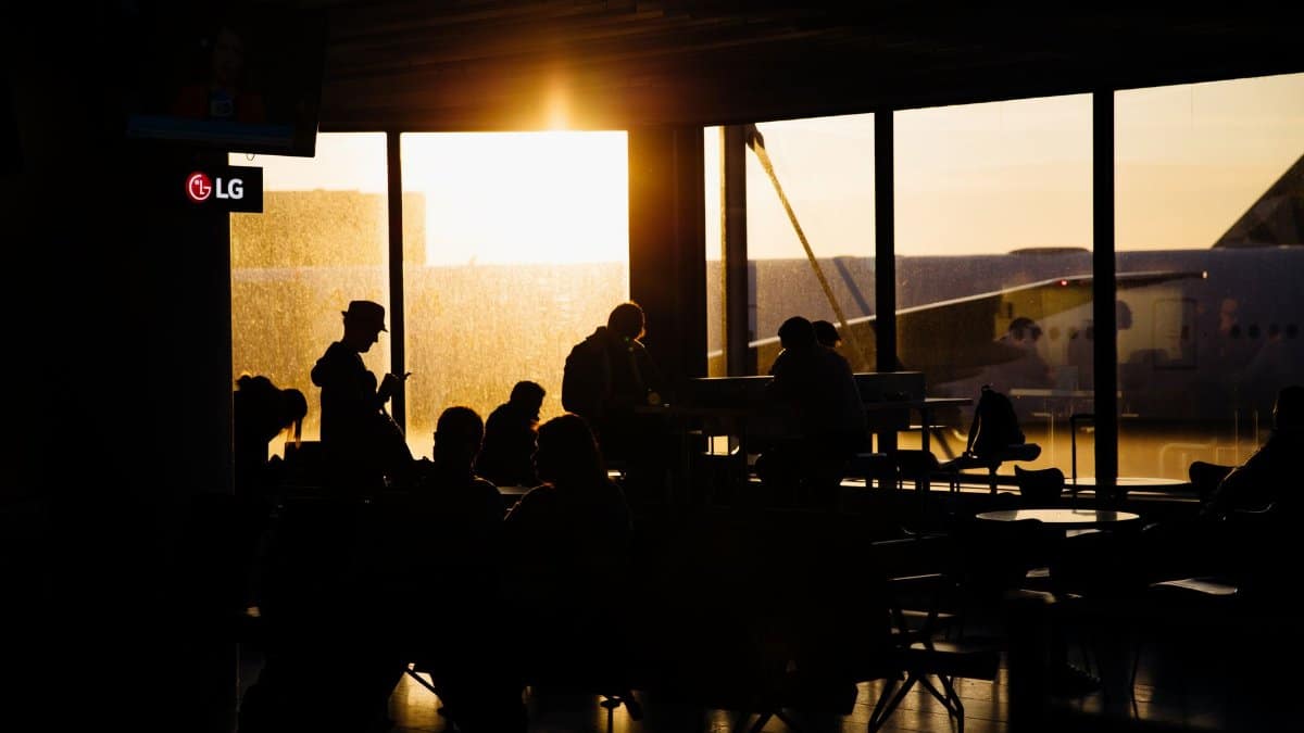 Travelers sitting in an airport terminal at sunset, silhouetted against the backdrop of an airplane.