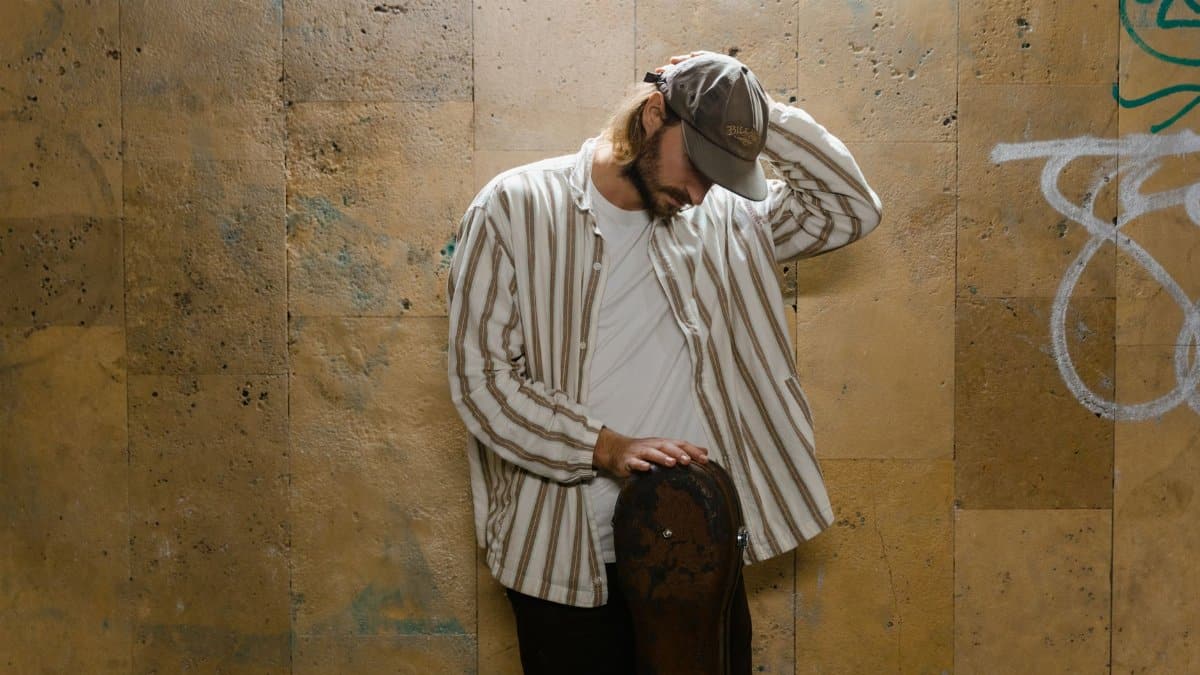 Man in casual attire leaning against a wall with a guitar case, evoking a contemplative street musician vibe.