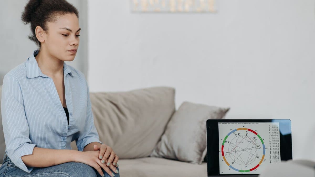 Young woman sitting in a living room analyzing an astrology chart on a laptop, reflecting concentration.