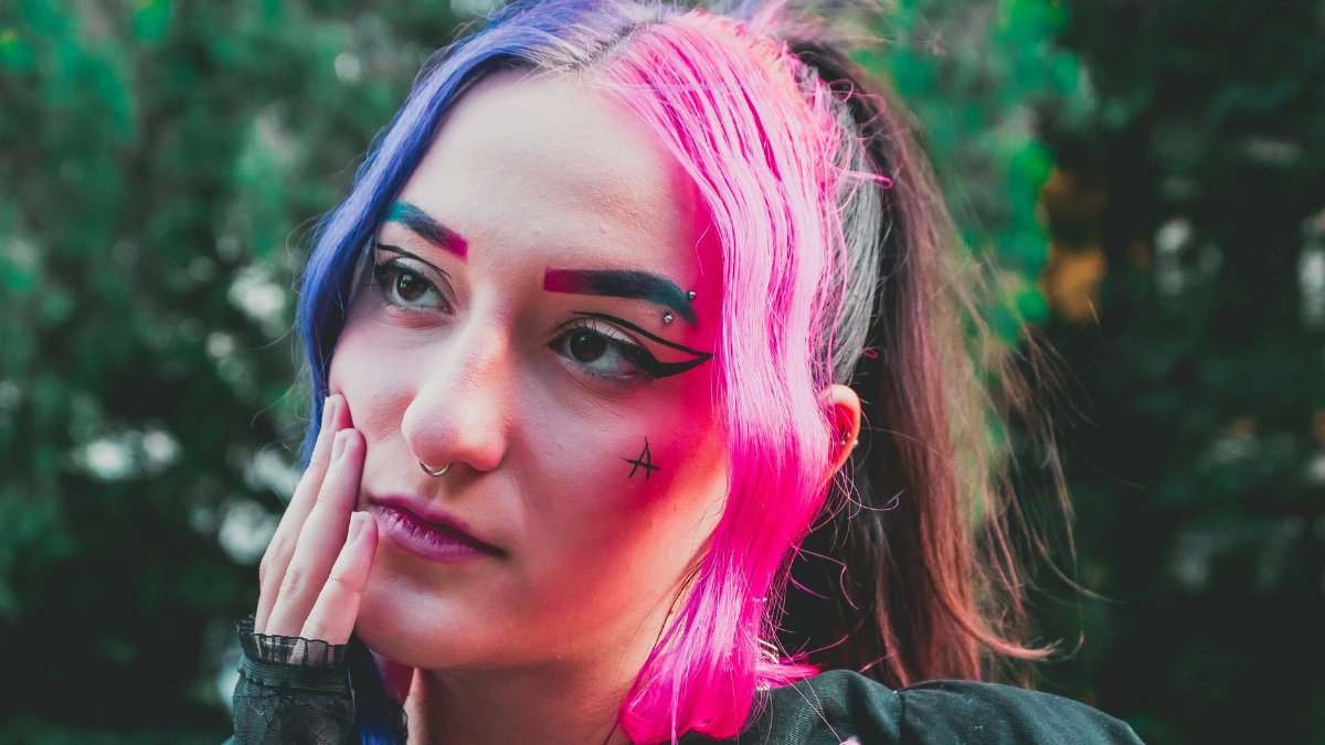 Young woman with vibrant colored hair and artistic makeup posing outdoors.