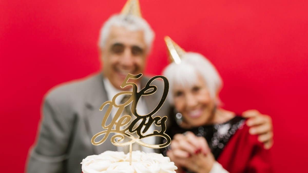 Elderly couple celebrating their golden anniversary with cake against a red background.