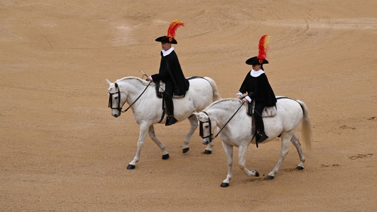 Two equestrians on white horses in traditional attire at Madrid arena.