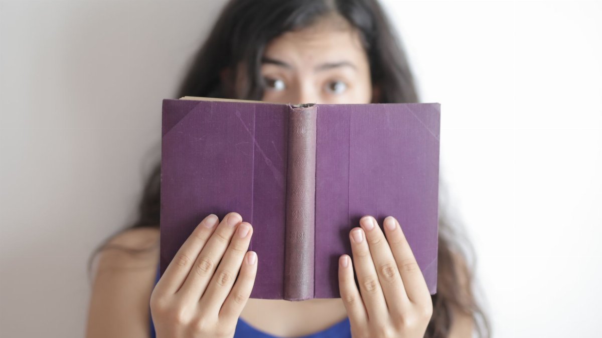 Blurred young woman in casual clothes standing against white wall while holding purple book in soft focus