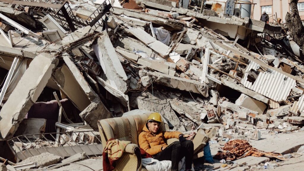 Young man in helmet on a sofa surrounded by destroyed buildings after earthquake.