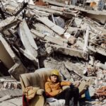 Young man in helmet on a sofa surrounded by destroyed buildings after earthquake.