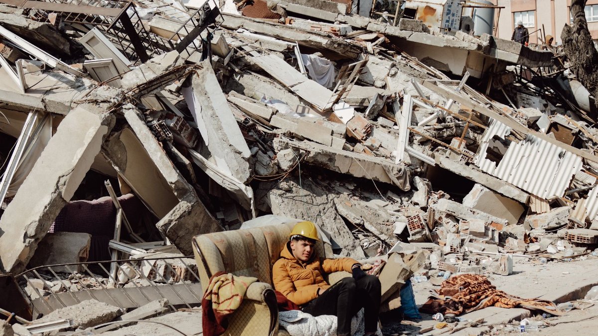 Young man in helmet on a sofa surrounded by destroyed buildings after earthquake.