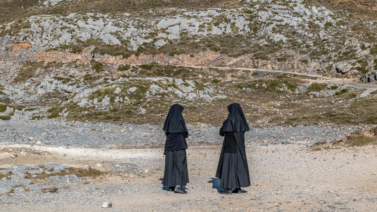 Two nuns in black habits walk in a rocky, remote landscape on a sunny day.