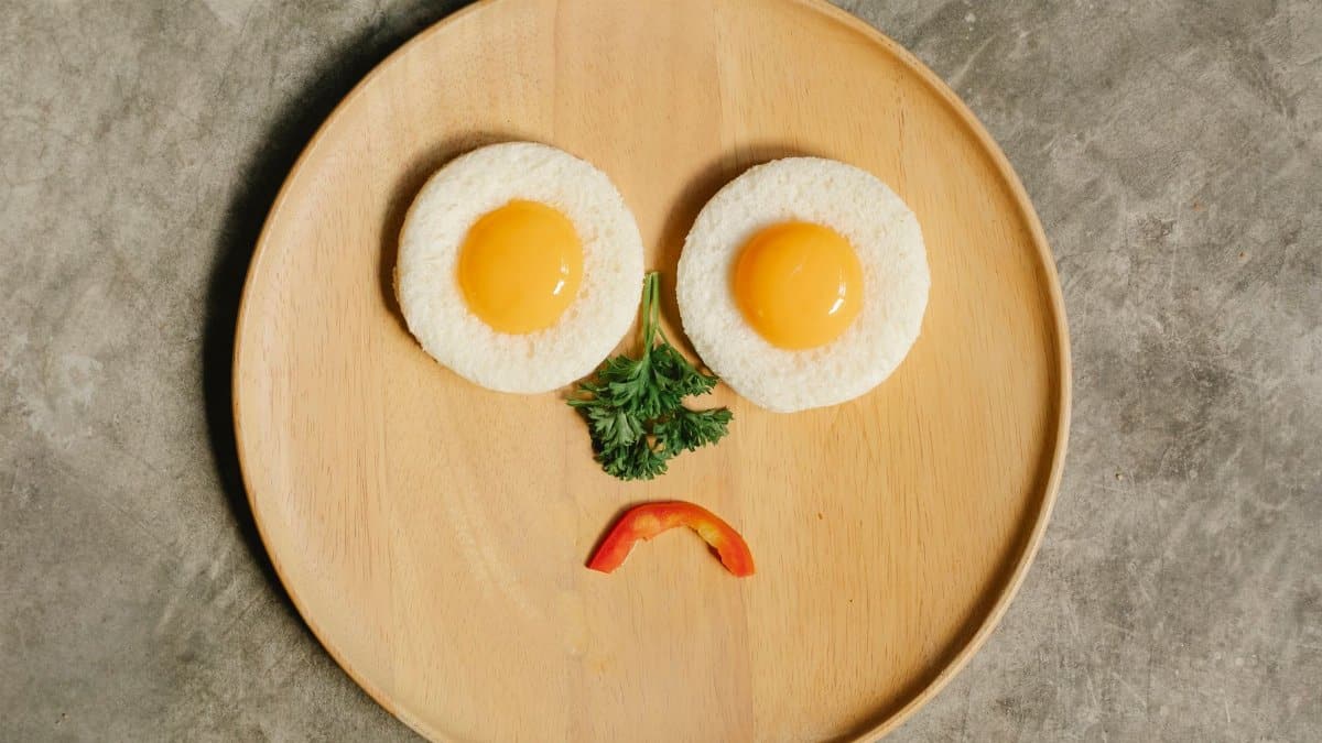 Top view of upset face made out of fresh yolks on round bread pieces with green parsley and bell pepper on wooden plate during breakfast