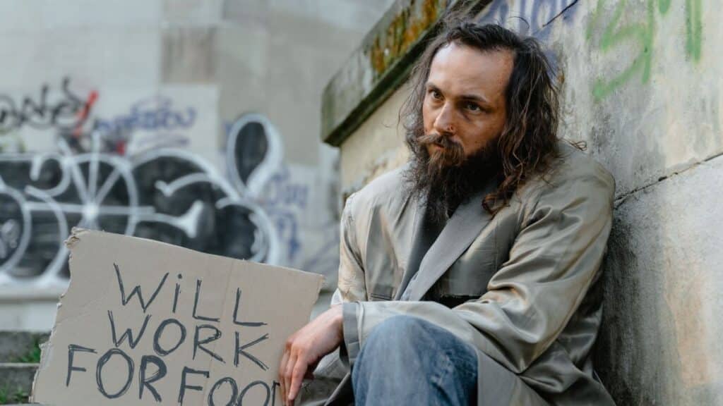 A homeless man with a sign expressing his willingness to work for food, sitting in an urban setting.