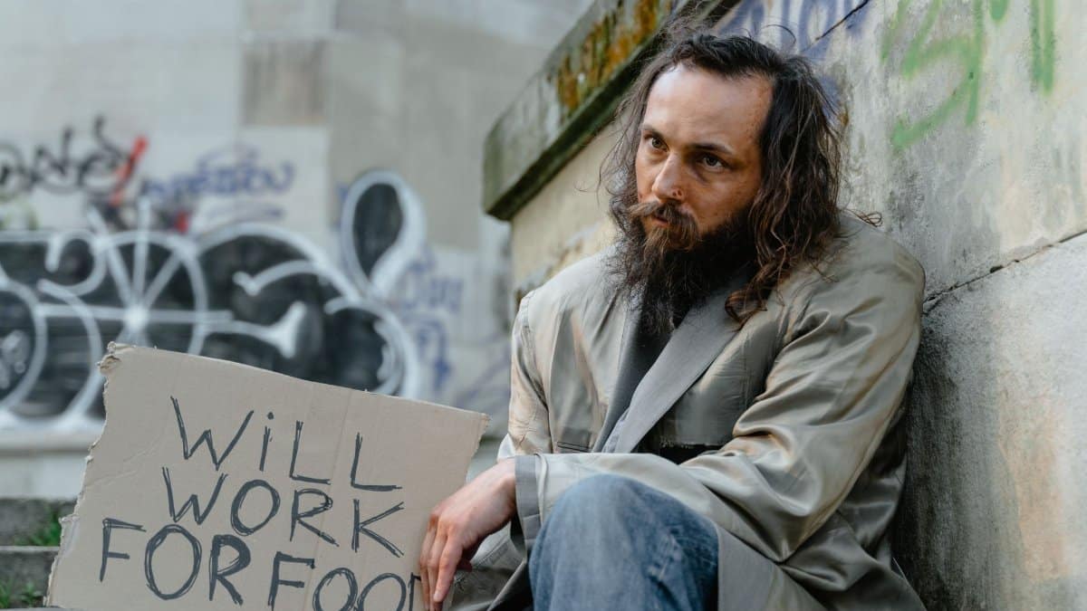 A homeless man with a sign expressing his willingness to work for food, sitting in an urban setting.