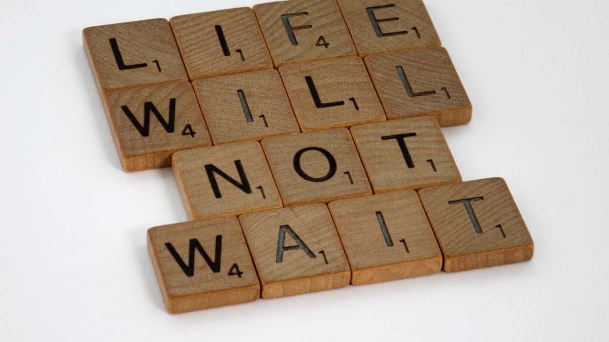 Wooden Scrabble tiles spelling 'Life Will Not Wait' on a white background.
