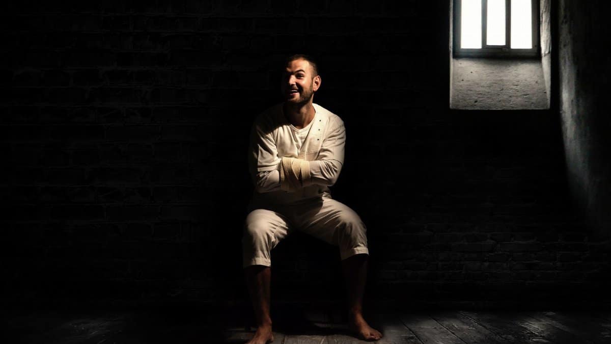 A man in a dimly lit room wearing a straightjacket, sitting with a smile in an asylum setting.