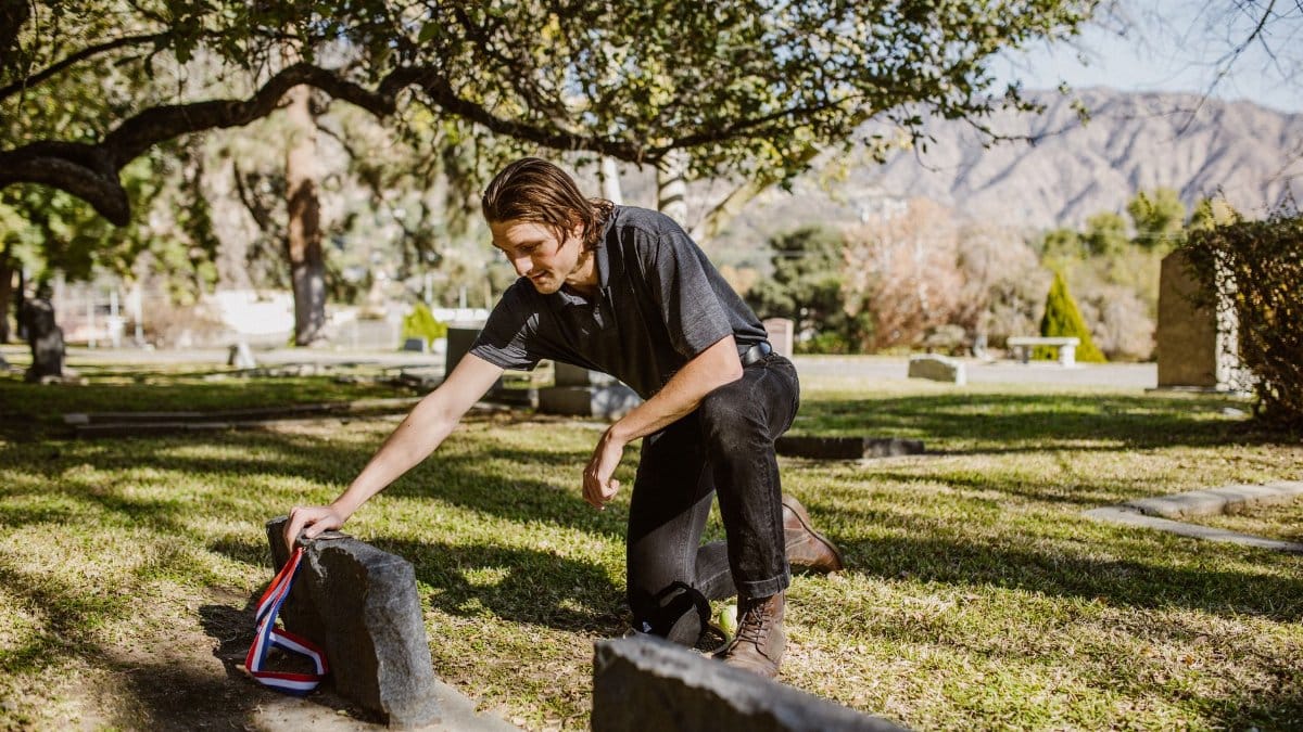 A man kneels beside a tombstone in a peaceful cemetery setting, symbolizing grief and remembrance.