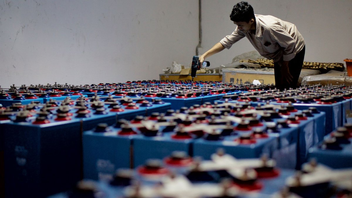 A worker checking many industrial batteries inside a facility. Indoor, industrial setting.