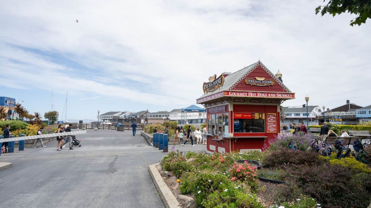 A colorful hot dog stand by the seafront promenade, surrounded by people enjoying a sunny day.