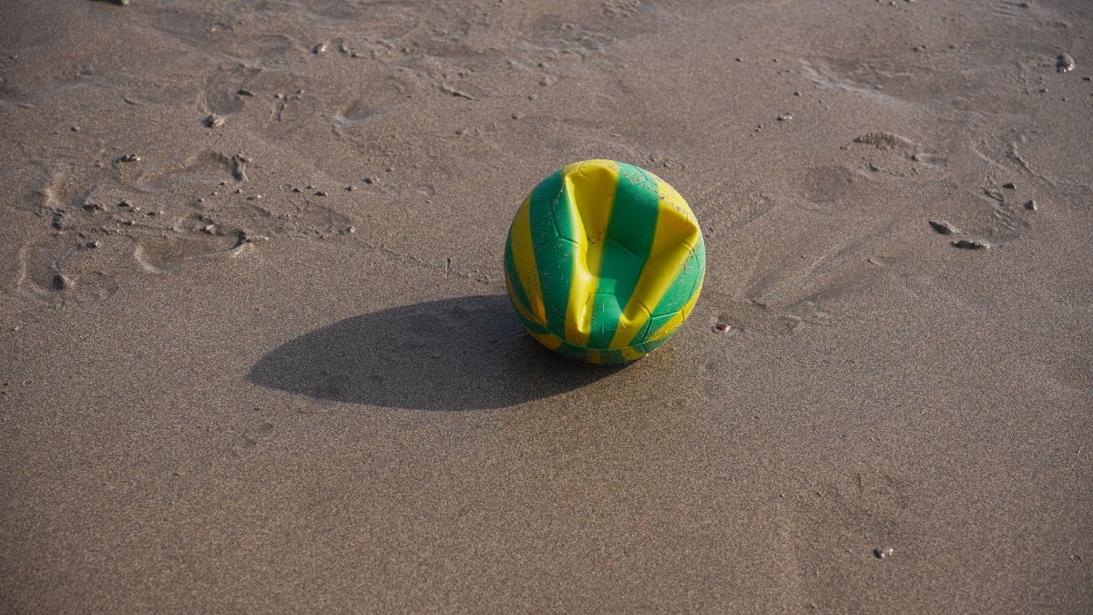 A deflated green and yellow ball resting on a sandy beach surface under bright daylight.