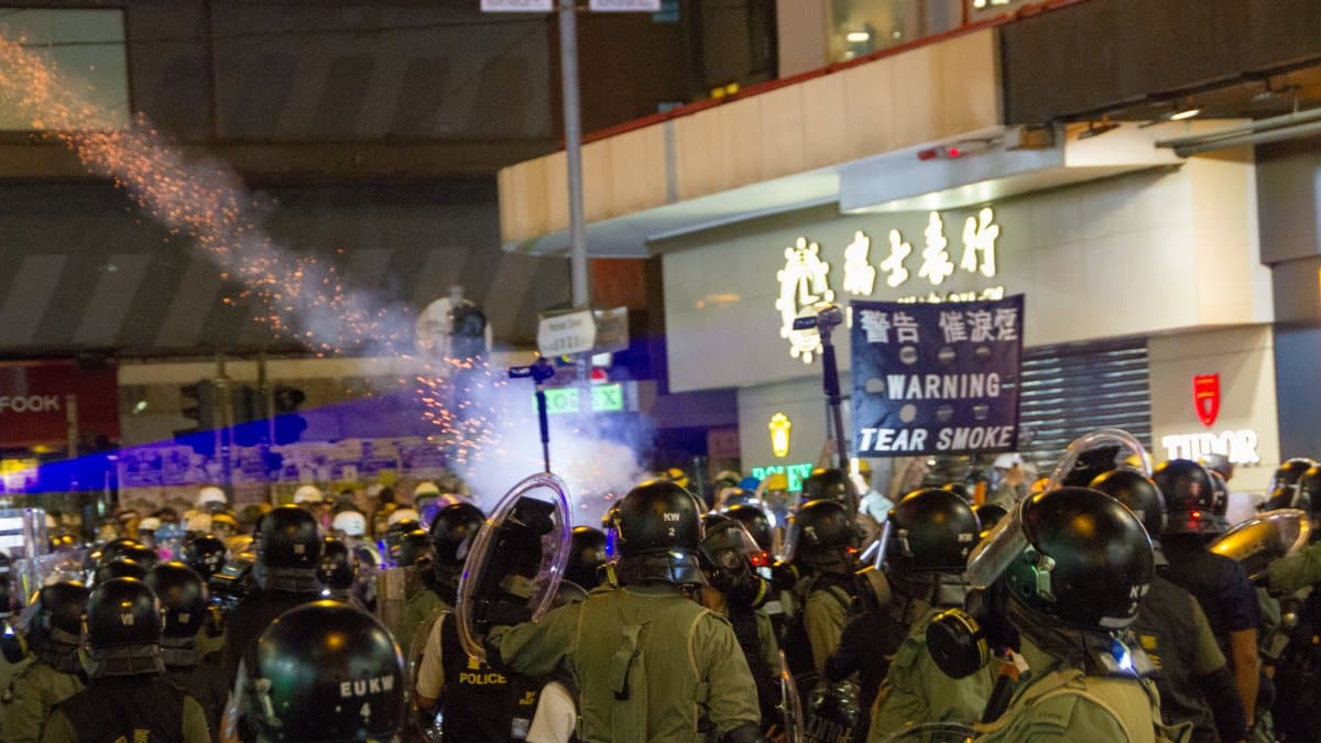 Riot police in tactical gear facing a public protest at night with tear gas.
