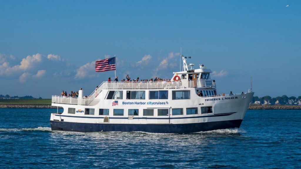A bustling Boston Harbor city cruise boat sails on a sunny day with an American flag waving prominently.