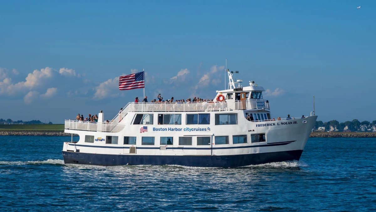 A bustling Boston Harbor city cruise boat sails on a sunny day with an American flag waving prominently.