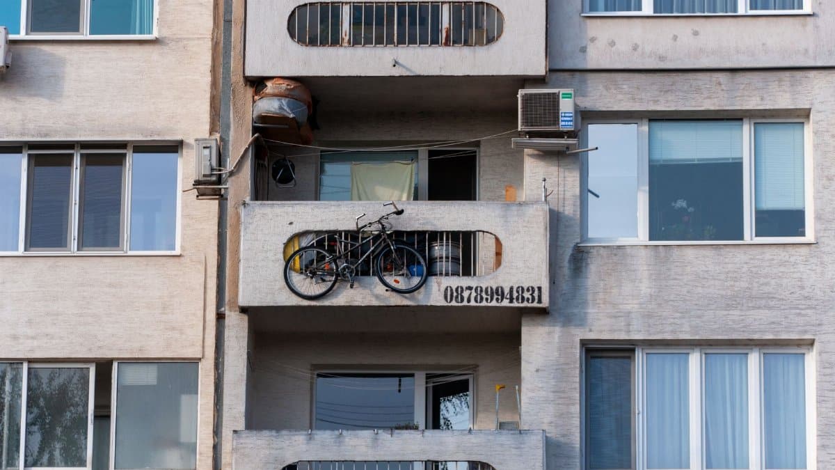 Urban scene of a concrete apartment building with a bicycle on the balcony and visible air conditioning unit.