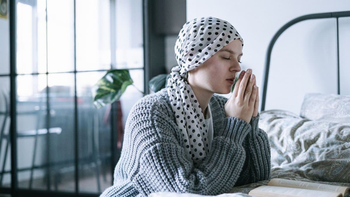 A hopeful woman in a scarf praying in a cozy bedroom during her cancer recovery journey.