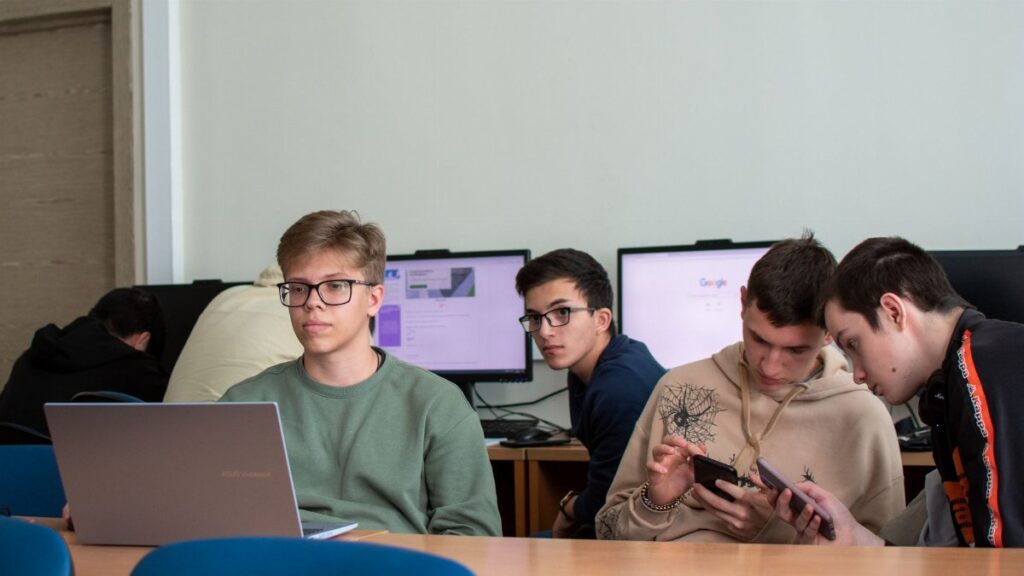 Five teenagers engaged with laptops and smartphones in a modern classroom.