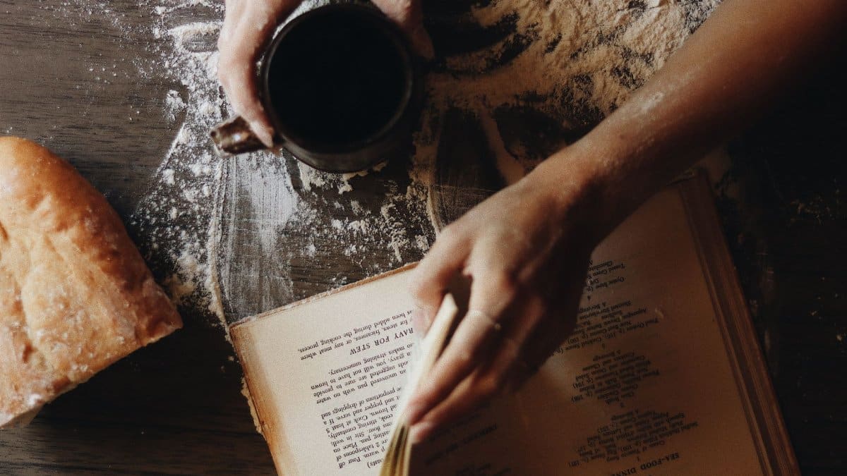 Hands with coffee cup and recipe book on flour-dusted table with bread.