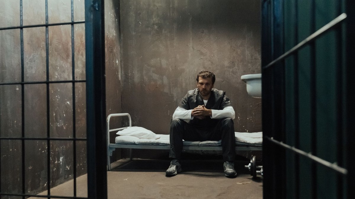 A man sits alone, reflecting inside a dimly lit prison cell.