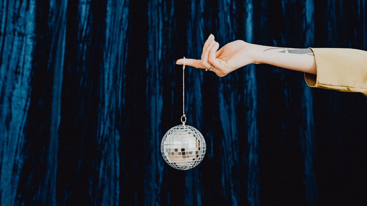 A stylish hand holds a sparkling disco ball against a dark blue curtain backdrop.