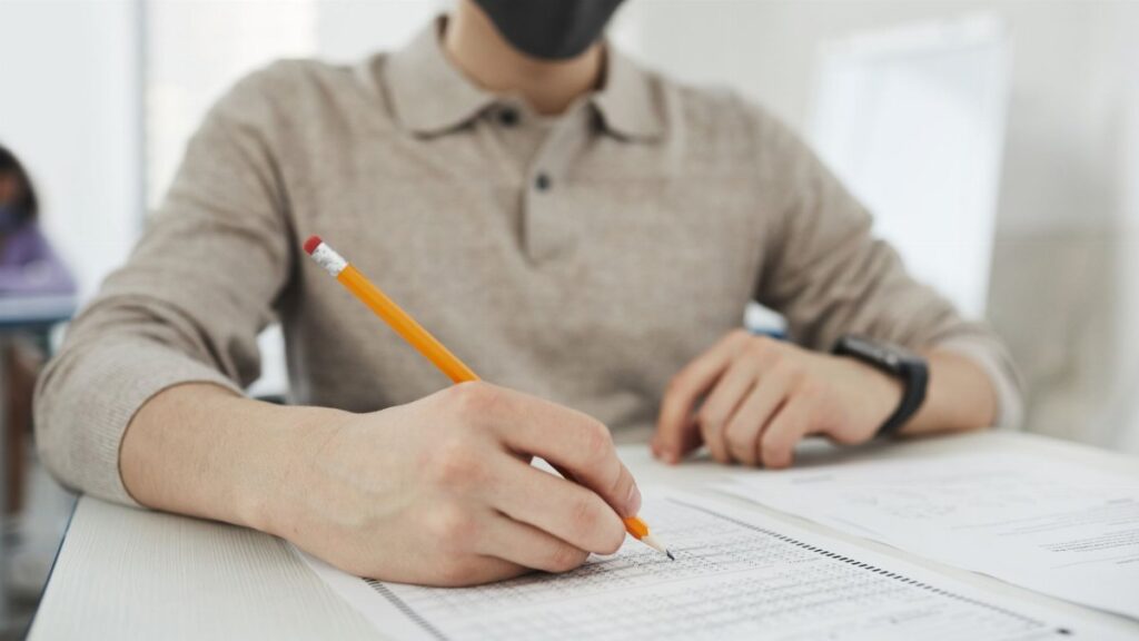 Person wearing a face mask writing a test at a desk, focusing on public health safety.