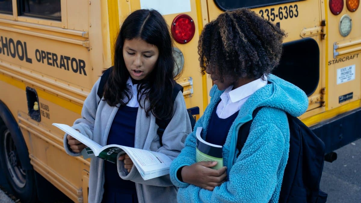 Concentrated multiethnic classmates holding textbooks and discussing study issues while standing near yellow school bus