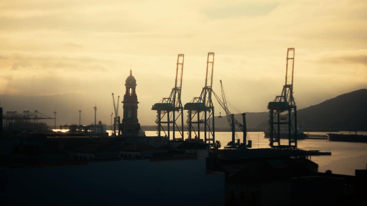 Silhouette of cranes and clock tower at sunset along a tranquil seaside port.
