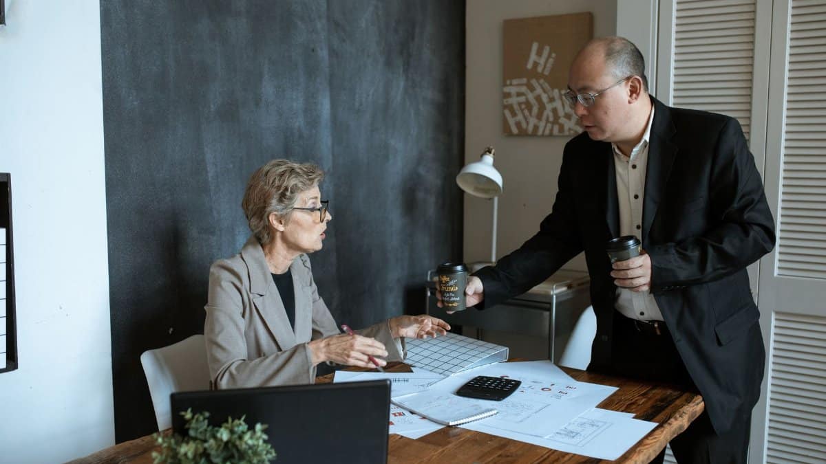 Two professionals in a modern office discussing work over coffee. Papers and laptop on desk.