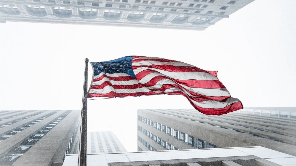 American flag waving amidst towering skyscrapers during a snowstorm in Chicago.
