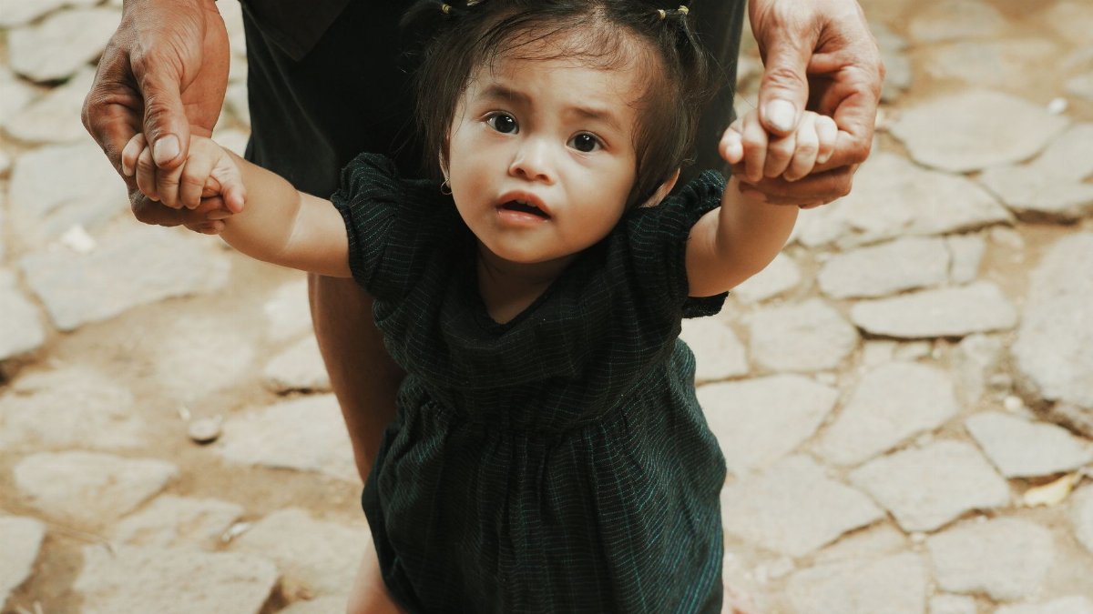 A cute baby girl takes her first steps while being held by an adult outdoors on a stone path in Banten, Indonesia.