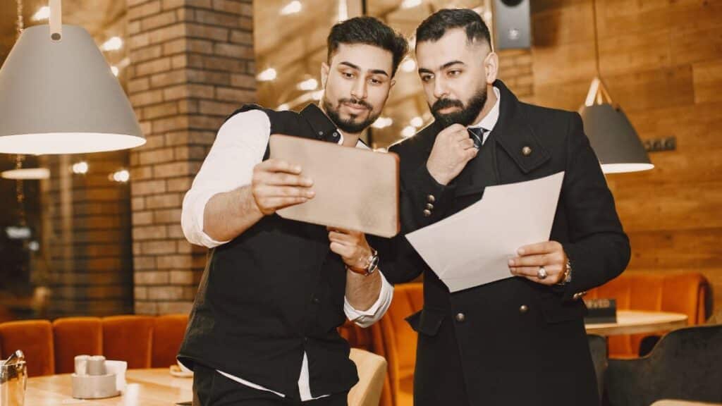 Two men in a restaurant review business documents and a tablet for planning purposes.