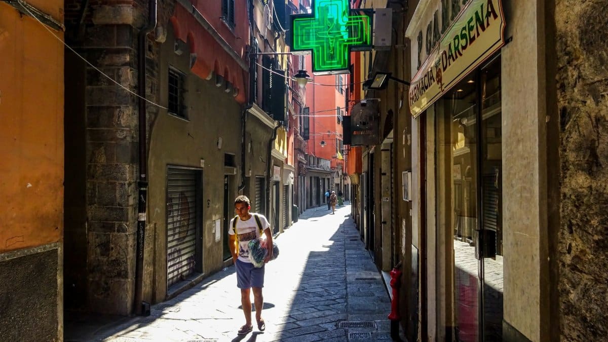 A local man walking through a narrow, sunlit alley in Genoa, Italy, under the iconic green pharmacy sign.