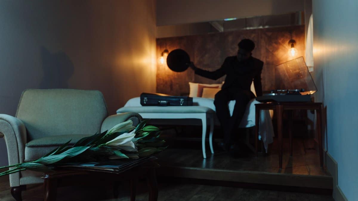A man in a dimly lit room, seated beside a bed with lilies, holding a record.
