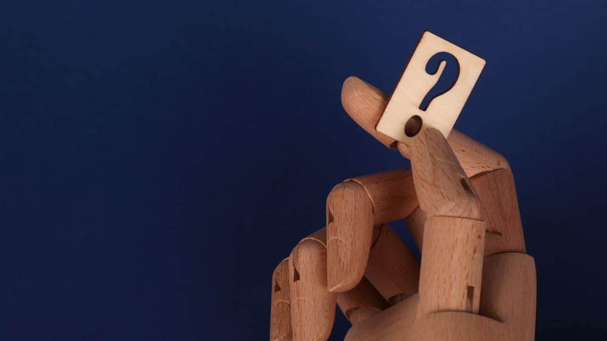 Wooden mannequin hand holding a block with a question mark against a blue background.