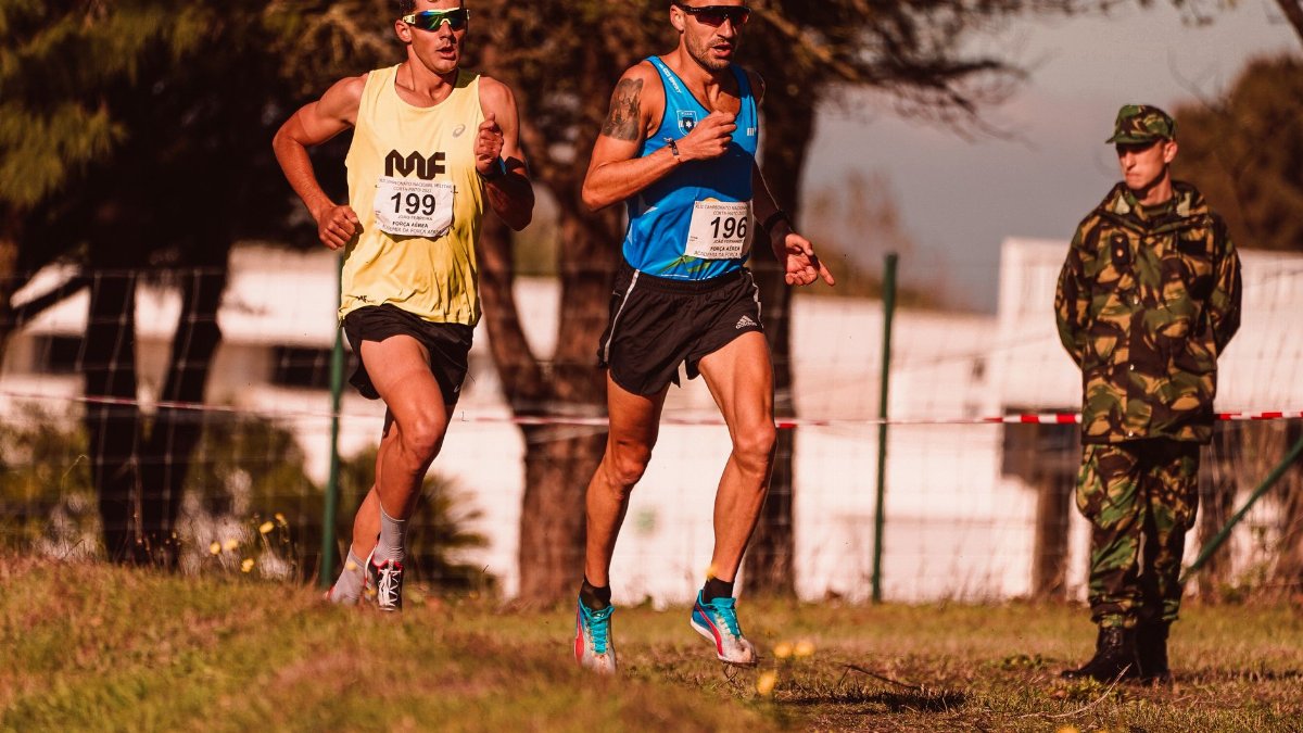 Two male athletes running outdoors wearing race bibs and sportswear, observed by a uniformed official.