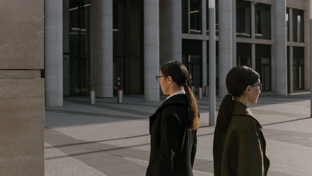 Two professional women in coats walking near a modern urban office building during the day.