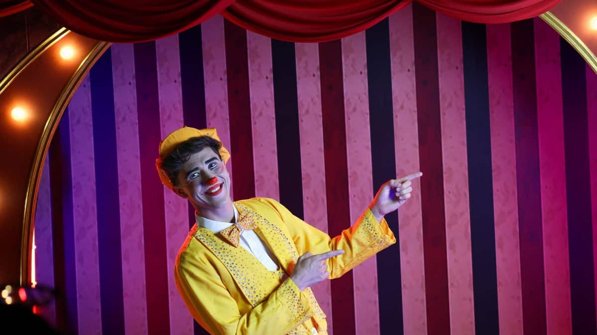 Cheerful clown in a vibrant yellow suit pointing during a stage performance, against a striped background.