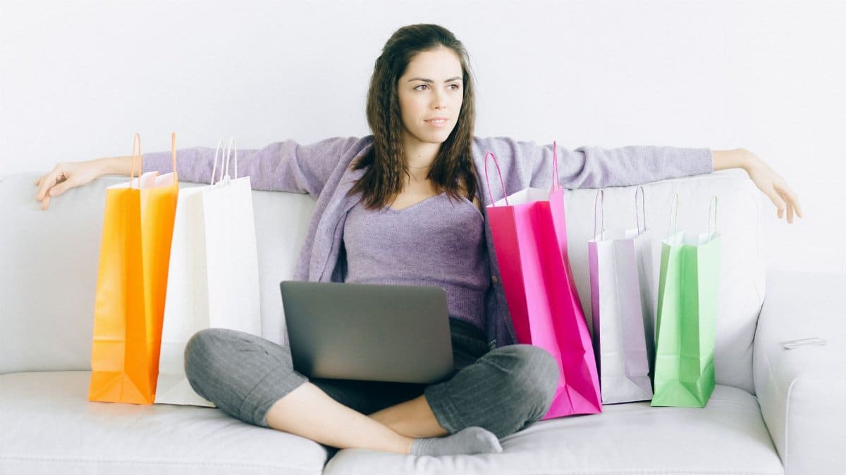 Young woman sitting on a couch with colorful shopping bags and a laptop, representing online shopping.