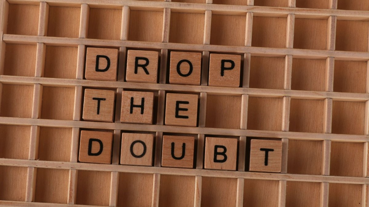 Wooden letter tiles spelling 'Drop the Doubt' on a wooden grid background, promoting positivity.