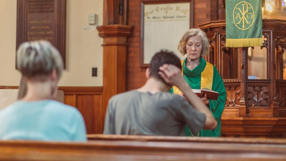 An elderly female clergyperson leads a service inside a church, engaging with the congregation.